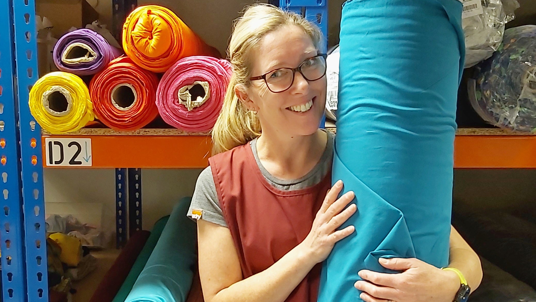 Mady standing in front of fabric rolls on shelves while holding a teal roll. She is smiling at the camera