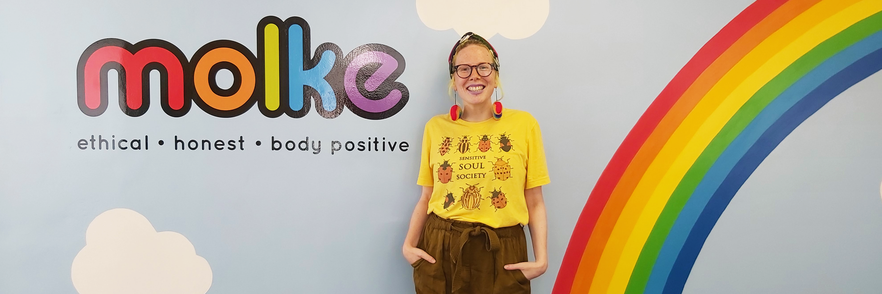 Jennifer stands in front of a light blue wall with a rainbow and the Molke logo painted on it. 