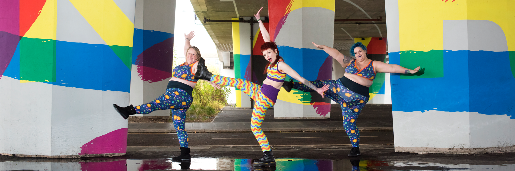 Three models are wearing molke bras and leggings and kicking to the left with their arms out, smiling.