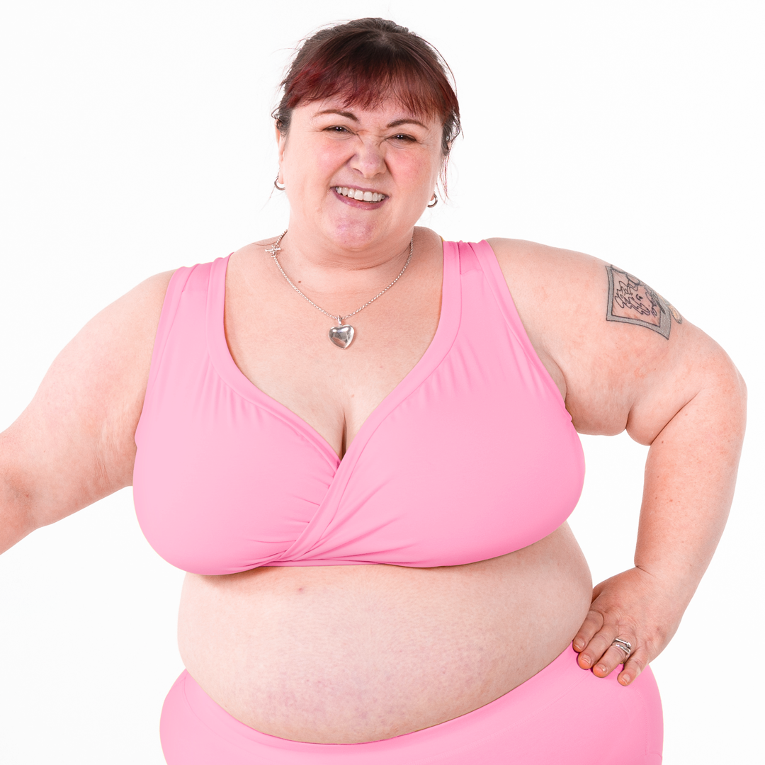 A smiling woman wearing a pink wrap-style non wired bra and matching pink bottoms, posed against a plain white background.