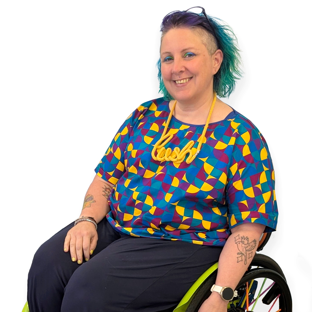 Kirsty is sitting in their wheelchair against a white background wearing a boxy cotton t-shirt in a busy geometric pattern.