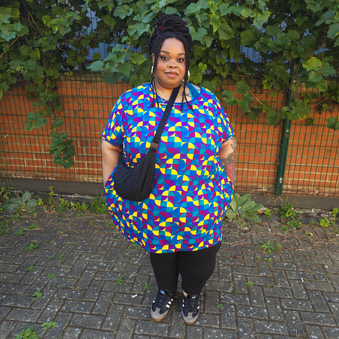 A woman stands outdoors on a paved walkway in front of a fence and green foliage, wearing an oversized short-sleeve top with a bold geometric multicolor pattern in blue, red, yellow, and purple, paired with black pants and a black shoulder bag.
