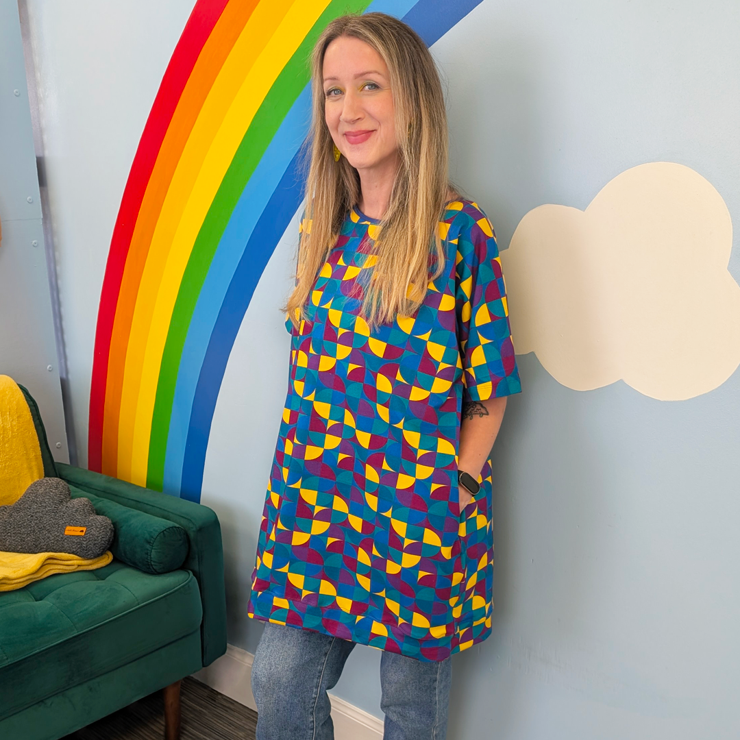Ros wearing a tunic dress with jeans leaning against a rainbow mural.