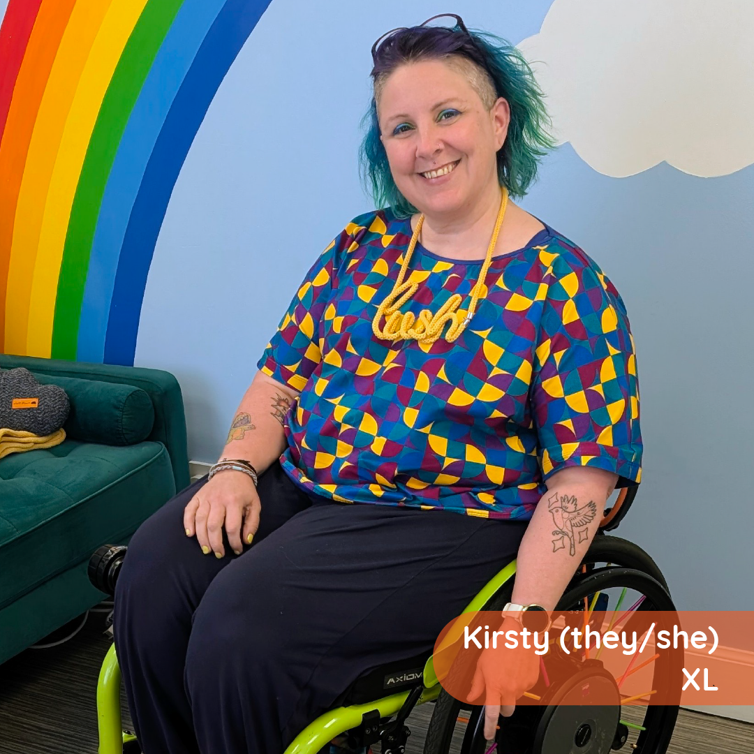 Kirsty in their wheelchair wearing a boxy tee in Eclipse in a room with a green sofa and a rainbow mural.