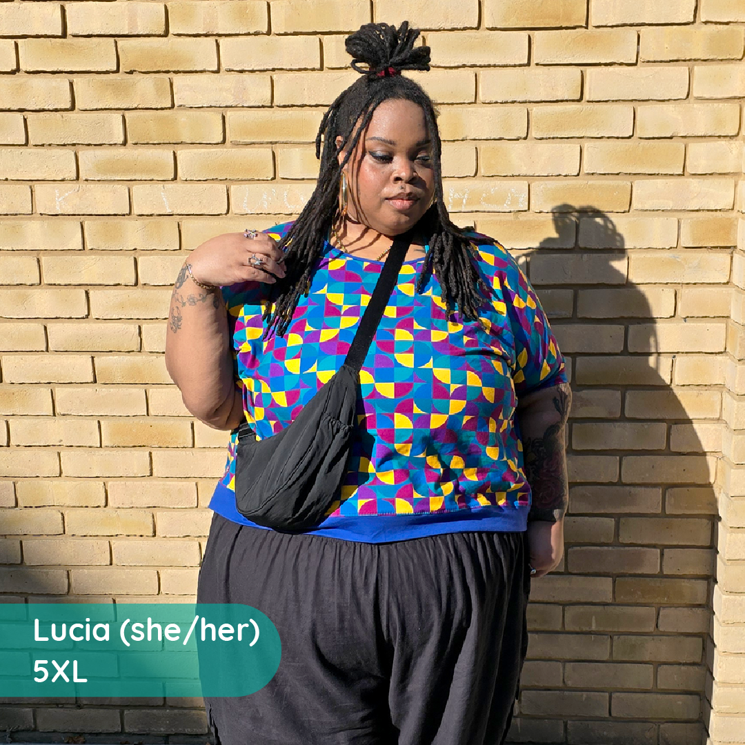 A plus-size woman stands against a tan brick wall wearing a colorful geometric print top with a blue base and purple, pink, and yellow shapes, dark gray pants, and a black crossbody bag. A teal text bubble in the lower left reads 'Lucia (she/her) 5XL'.
