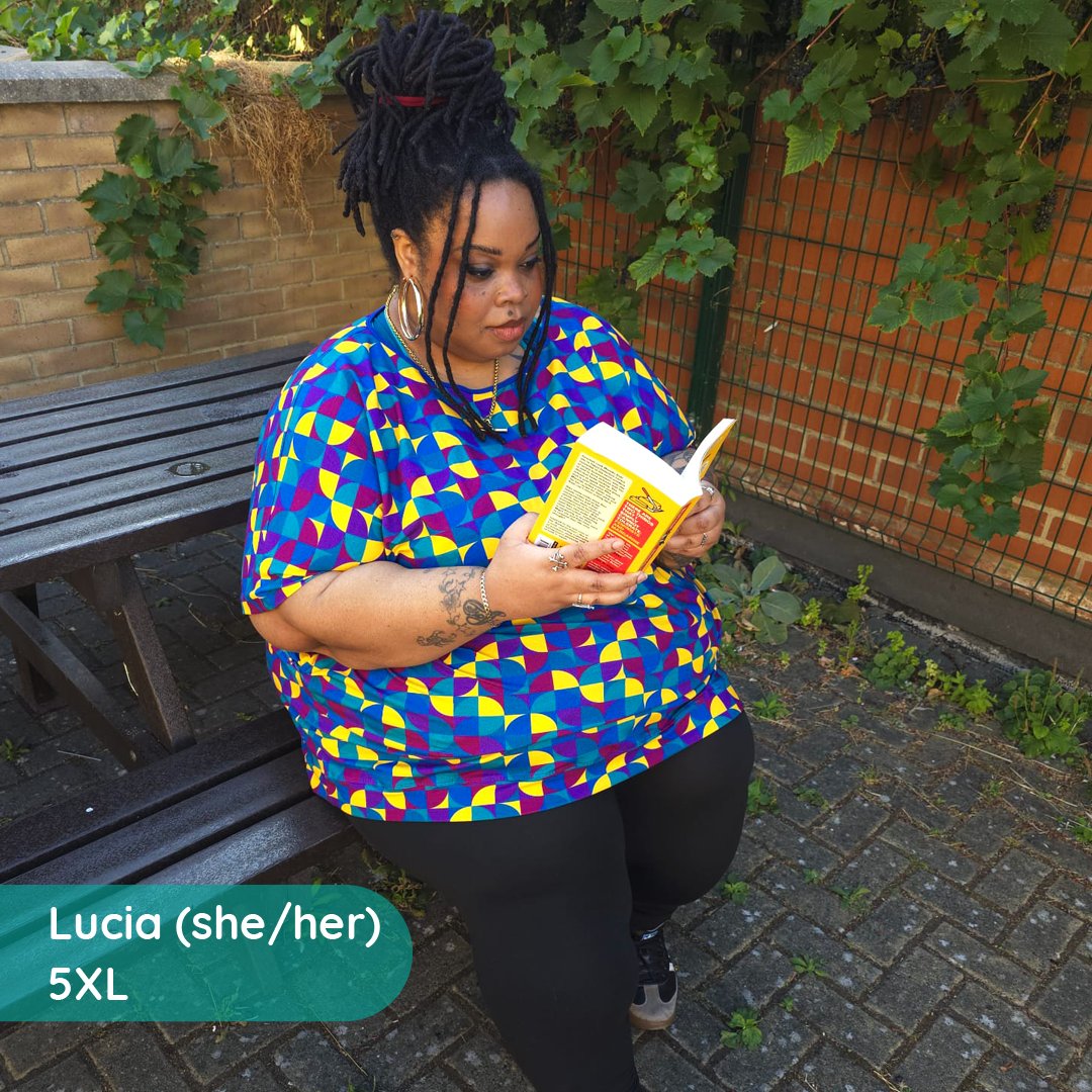 A woman sitting on a wooden bench outdoors, reading a book. She is wearing a bright short-sleeve t-shirt with a bold geometric pattern in blue, purple, yellow, and red, paired with black leggings.