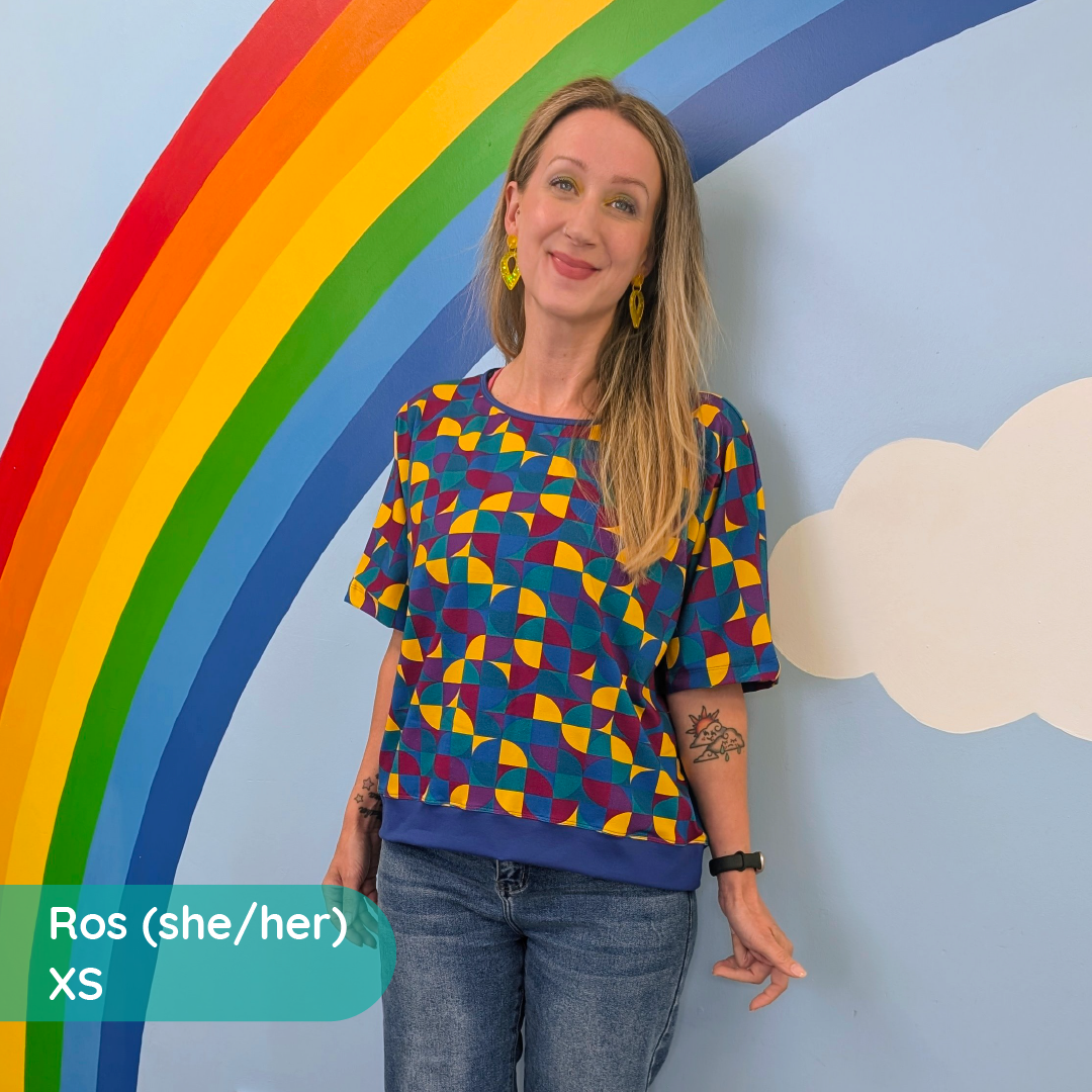 Ros is standing against a wall with a rainbow painted on it wearing a boxy tee in Eclipse and jeans.