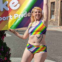Eilidh is wearing a Pride rainbow bralette and mid rise briefs holding up a Pride banner