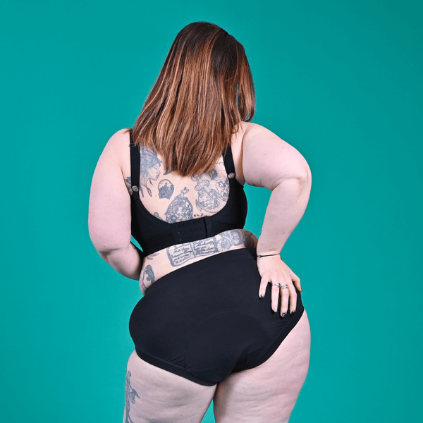 Back view of Kat wearing black high rise period pants and a flexi size black bra against a teal background